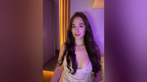 Media: Video of a young woman with long, wavy brown hair, wearing a white lace lingerie top, standing indoors with purple lighting.