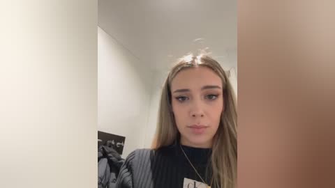 Media: Video of a young Caucasian woman with long, straight blonde hair, wearing a black top and a lanyard, standing in a dimly lit hallway with white walls and a framed photo.