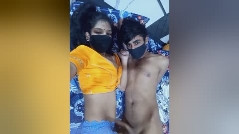 Media: Video of two topless South Asian individuals lying on a blue and white floral bedspread, both wearing black masks.