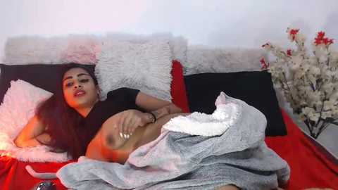 Media: Video of a young woman with long dark hair, medium skin tone, and slender physique, lying on a red bed, partially covered by a gray blanket, wearing a black top.