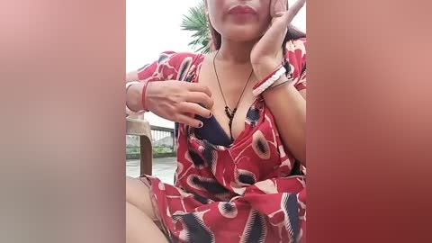 Media: A video of a light-skinned woman with long hair, wearing a red dress with black and white geometric patterns, holding a blue phone against her chest, seated outdoors.