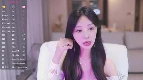 Media: A video of an Asian woman with long black hair, wearing a white off-shoulder top and dangling earrings, seated indoors.