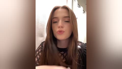 Media: Video of a fair-skinned young woman with long, straight, light brown hair, wearing a black, lacy top, pouting in a bathroom with white tiled walls and a mirror.