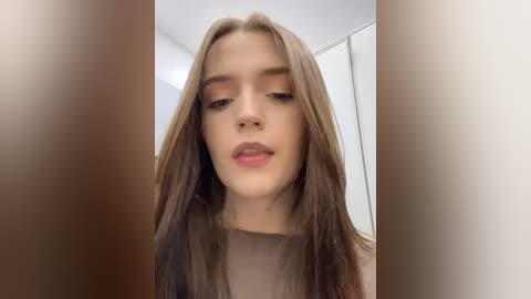 Media: A close-up video of a young woman with long, straight brown hair, fair skin, and natural makeup, looking directly at the camera in a neutral expression. The background is a simple, off-white room with soft lighting.