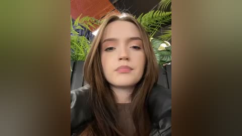 Media: Video of a young woman with straight brown hair, wearing a black leather jacket, standing in front of lush green plants and orange wall art.