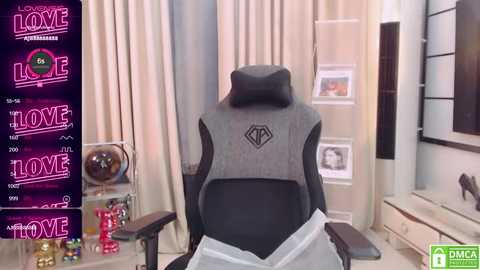 Media: A video of a gaming chair with a black headrest and gray backrest, set in a cozy room with beige curtains, framed photos, and a TV. \"Love Home\" logo on the left.