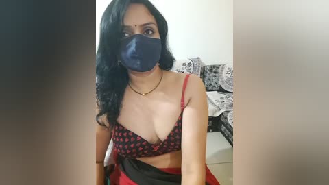 Media: Video of a South Asian woman with long black hair, wearing a black face mask, red saree, and a black bra with red heart pattern, seated indoors.