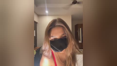 Media: Video of a young woman with long, light brown hair, wearing a black face mask, red tank top, and standing in a dimly lit, beige-walled room with a ceiling fan and open door visible in the background.