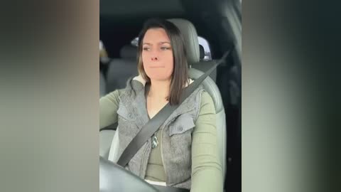 Media: Video of a woman with shoulder-length brown hair, wearing a gray puffy vest, driving a car with a seatbelt on. The background shows the car interior.