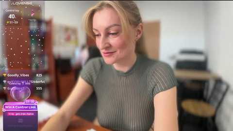 Media: Video of a smiling, light-skinned woman with blonde hair, wearing a green mesh top, seated in a room with a wooden desk, bookshelf, and printer.