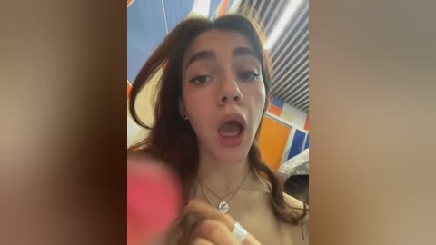 Media: Video of a young woman with long, brown hair, wearing a necklace, making a suggestive face in a bathroom with tiled walls and a fluorescent light.