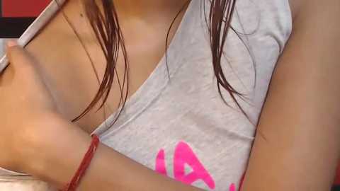 Media: Close-up video of a young woman with wet, dark brown hair, wearing a grey tank top with pink writing, and a red bracelet on her wrist.