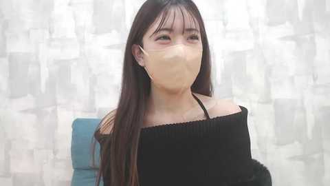 Media: Video of an Asian woman with long, straight dark hair, wearing a black off-shoulder top, and a beige face mask. She stands against a patterned white and gray background.