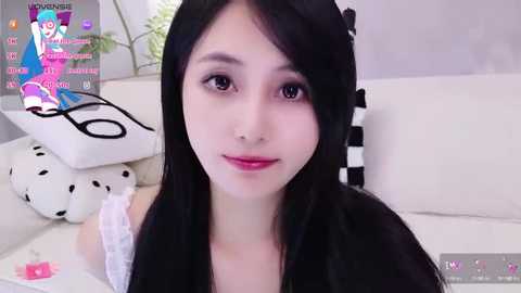 Media: A young Asian woman with long black hair, wearing a black dress, sits on a white couch with a black and white pillow. The background features a green plant and a \"Minecraft\" video game overlay.