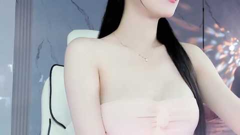 Media: Video of a slender East Asian woman with long black hair, wearing a strapless white top, seated in a modern, minimalist room with gray walls and a white chair.