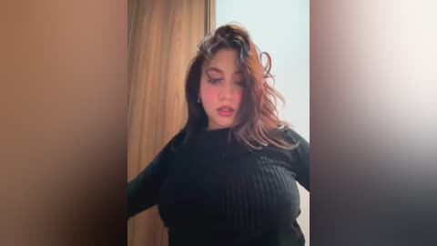 Media: Video of a young woman with light brown skin, wearing a black ribbed sweater, standing indoors with a blurred background.