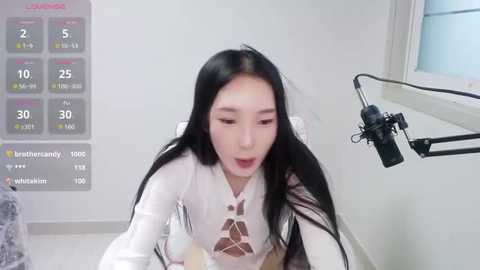 Media: A video of a young East Asian woman with long black hair, wearing a sheer white top with lace cutouts, singing into a microphone in a minimalist studio.