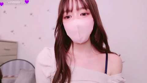 Media: Video of an Asian woman with long brown hair, wearing a white face mask and a white off-shoulder top, standing in a room with white walls and a fan.