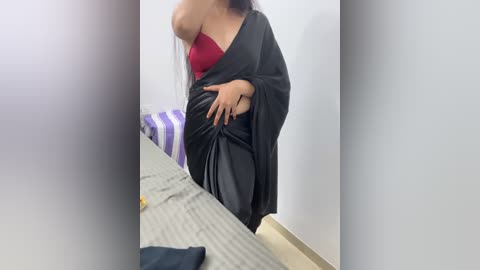 Media: A video of a woman with medium brown skin, wearing a red bra and black sari, standing in a bedroom with a striped purple and white bedspread and grey sheets.