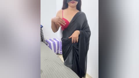 Media: Video of a woman with long black hair, wearing a red bra and black sari, standing in a minimalist room with a striped bed and white walls.