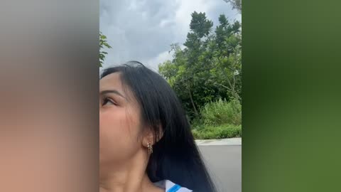 Media: Video of a young Asian woman with long black hair, wearing earrings, captured from the side, with blurred background showing green foliage and a cloudy sky.