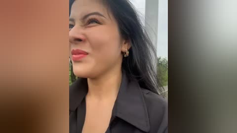 Media: A video of a woman with long black hair, light skin, and a medium build, wearing a black blazer. She has a slight smile and wears small gold earrings. The background shows blurred trees.