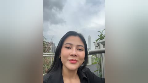 Media: Video of a young Asian woman with long black hair, wearing a black jacket, standing on a balcony with a cloudy sky and greenery in the background.