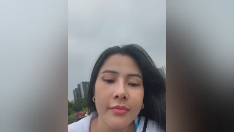 Media: A video of a young East Asian woman with straight black hair, wearing a white jacket, looking down thoughtfully. Background shows blurred buildings and greenery, likely taken outdoors.