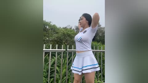 Media: Video of a young Asian woman in a white cheerleading outfit, with a short skirt and black trim, stretching outside a green fenced garden.