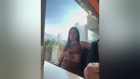 Media: A video captures a woman with long, dark hair and medium skin tone, wearing a brown top, sitting inside a modern building with large windows and a city skyline visible outside.