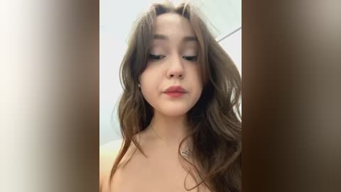 Media: Video of a young woman with long, wavy brown hair, fair skin, and light makeup, topless, looking down with a neutral expression. Background is blurred.