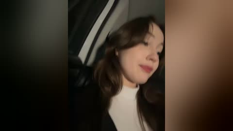 Media: Video of a young woman with long brown hair and fair skin, wearing a white top, seated in a car with black leather seats. She has a soft smile, and the background is dimly lit.