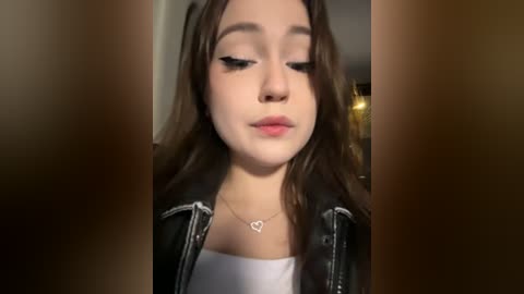 Media: Video of a young woman with fair skin, brown hair, wearing black leather jacket, white top, and heart necklace, looking down, blurry background.