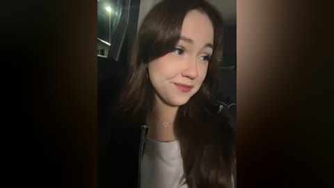 Media: Video of a young woman with long brown hair, wearing a black jacket over a white top, smiling subtly. Background features a dimly lit room with a window and a faint reflection.