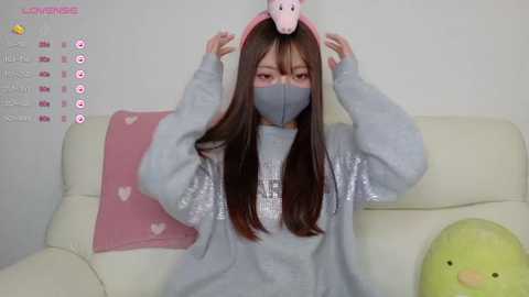 Media: A video of a young Asian woman in a gray mask, light blue sweater, and pink bunny ears, sitting on a cream-colored couch with a pink pillow.