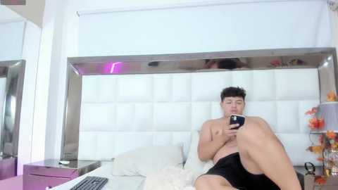 Media: Video of a shirtless Asian man in black shorts sitting on a white bed, holding a smartphone, in a modern, white-themed room with a large, reflective wall.