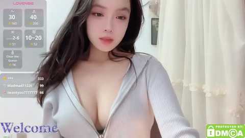 Media: Video of an East Asian woman with long black hair, fair skin, and large breasts, wearing a low-cut gray sweater, in a softly lit bedroom.