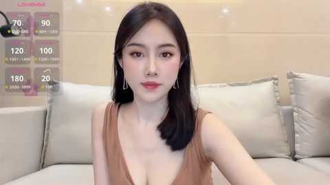 Media: Video of a fair-skinned East Asian woman with straight black hair, wearing a brown sleeveless dress, sitting on a beige sofa. Background includes a modern, minimalist living room with cream-colored walls and cushions.