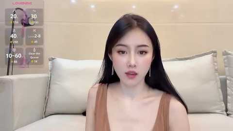 Media: Video of an Asian woman with long black hair, fair skin, and subtle makeup, wearing a sleeveless brown dress, sitting on a white sofa with cream pillows in a modern living room.