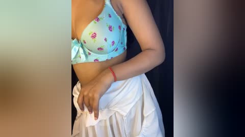 Media: Video of a woman with medium brown skin, wearing a light blue bra with pink floral patterns and a white skirt, standing against a dark background.