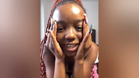 Media: Video of a smiling young Black woman with dark skin, wearing a red headwrap and pink top, hands framing her face, with a blurred indoor background.