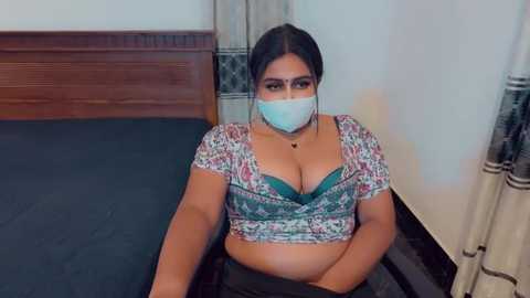 Media: A video of a curvy woman with dark skin, wearing a floral crop top, face mask, and black pants, sitting on a bed with a wooden headboard and patterned curtains in the background.