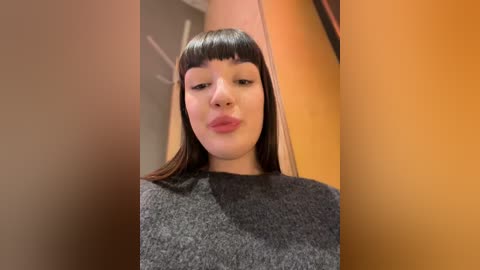 Media: Video of a young woman with straight, dark hair, blunt bangs, and fair skin, wearing a grey sweater, standing in an orange-toned room with blurred background.