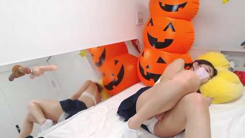 Media: Video of a woman in Halloween costume, lying on a bed, with a large jack-o'-lantern and yellow pillow, wearing a mask and dark clothing, white sheets, and a mirror reflecting her.