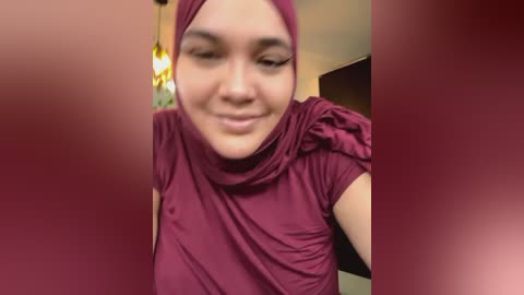 Media: Video of a smiling woman with a light brown complexion, wearing a burgundy hijab and matching top, taken indoors with blurred background, warm lighting, and a hint of a ceiling lamp.