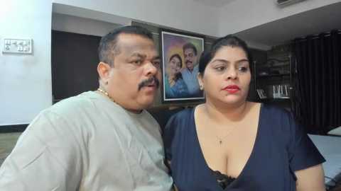 Media: Video of an overweight man with a mustache, wearing a beige shirt, and a plus-size woman with short black hair, red lipstick, and a deep V-neck blue blouse, both indoors against a dark wall with framed family photos.