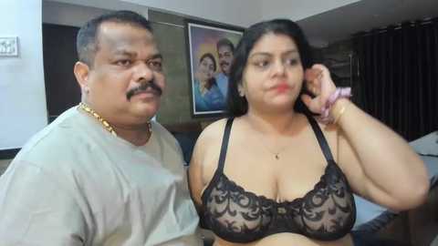 Media: Video of a middle-aged South Asian couple in a modest living room; the man wears a white T-shirt and gold chain, the woman a black lace bra. Background shows a framed picture of a couple on a wall, dark curtains, and a bed.