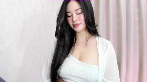 Media: Video of an Asian woman with long, black hair and fair skin, wearing a white, off-the-shoulder crop top, standing in a softly lit room with light-colored curtains.