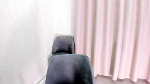 Media: A video of a minimalist room with a grey upholstered chair facing a white wall and a pink curtain on the right. The room has a soft, calming ambiance.