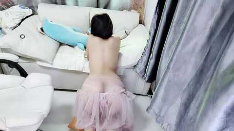 Media: Video of an Asian woman with black hair, kneeling nude on a white couch, wearing a pink tulle skirt. Background includes a white chair and gray curtains.